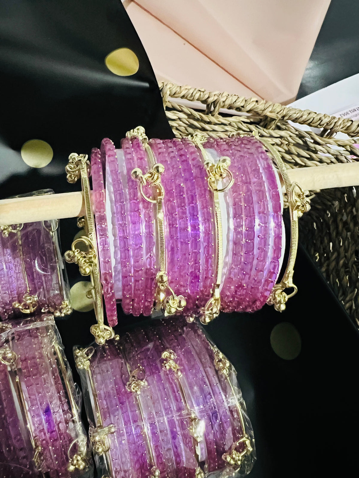 A close-up of multiple stacked purple beaded bangles with gold-toned metal connectors and decorative charms, arranged on a display stand with additional packaged bangles below.