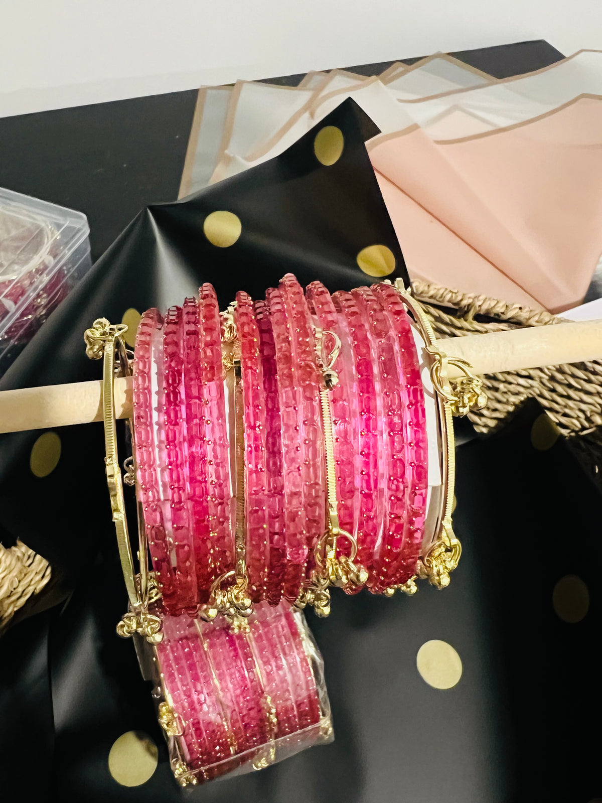 A set of pink beaded bangles with gold-toned metal borders and small dangling gold charms, displayed on a wooden stand with additional packaged bangles below, against a black and gold polka-dot background.