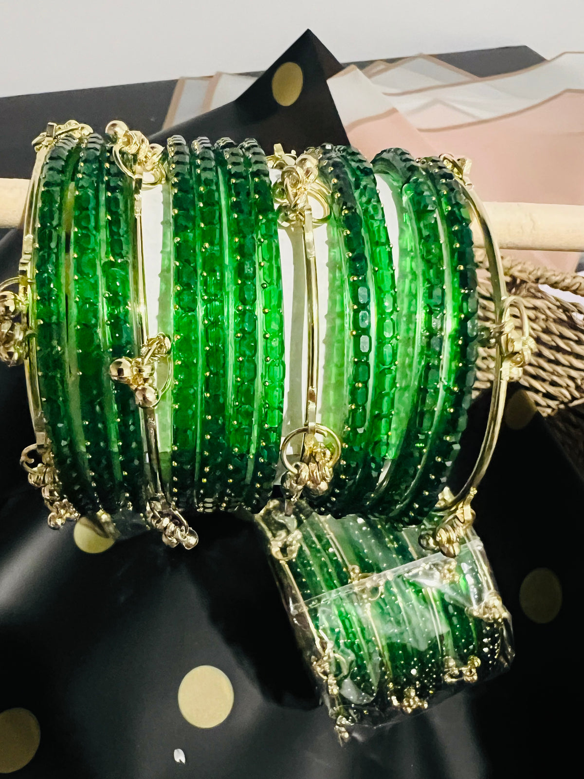 A stack of green beaded bangles with gold-tone metal accents and butterfly charms, displayed on a black polka-dot surface with a matching packaged set in front.