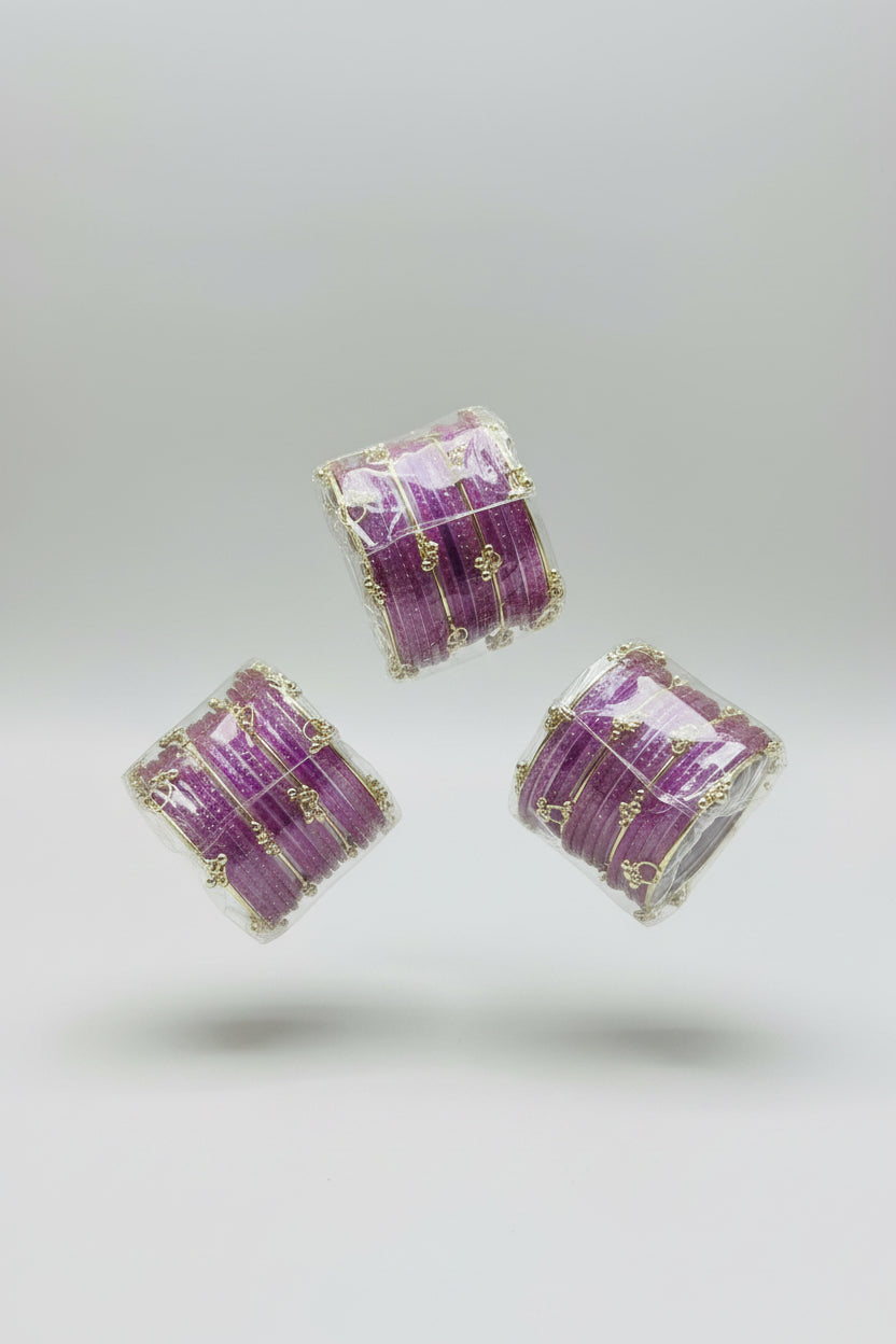 Purple glitter bangle sets with gold-plated bangles and small dangling charms, displayed on a wooden stick and in clear plastic packaging on a black polka-dot surface.