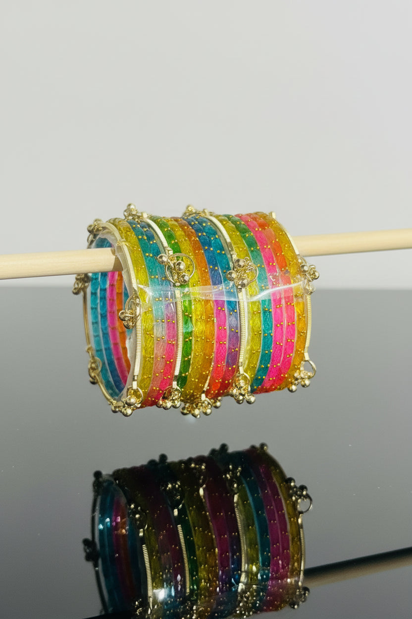 A close-up of a stack of colorful beaded bangles with gold-toned borders and small hanging charms, displayed on a wooden stick against a black and pink background.