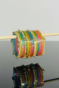 A close-up of a stack of colorful beaded bangles with gold-toned borders and small hanging charms, displayed on a wooden stick against a black and pink background.
