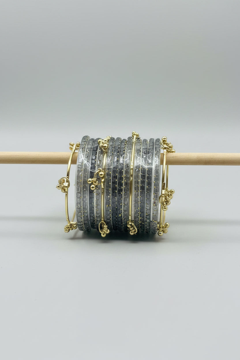 A stack of clear Glass bangles with gold-plated borders and decorative gold charms, wrapped in protective plastic and displayed on a wooden stand against a black polka-dot background.