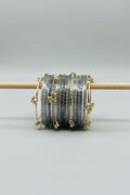 A stack of clear Glass bangles with gold-plated borders and decorative gold charms, wrapped in protective plastic and displayed on a wooden stand against a black polka-dot background.