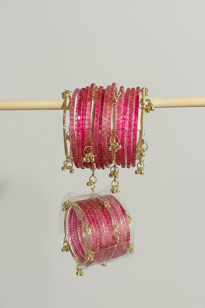 A stack of pink beaded bangles with gold-toned metal borders and dangling charms, displayed on a wooden stand with additional packaged bangles below, set against a black polka-dot surface.