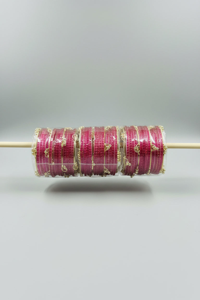  pink bangles with gold foil stripes and small gold decorative charms, wrapped in clear plastic and arranged on a wooden stick against a black background with gold polka dots.