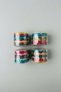 Four clear plastic-wrapped stacks of colorful bangles with gold-tone decorative elements, arranged on a woven straw surface.