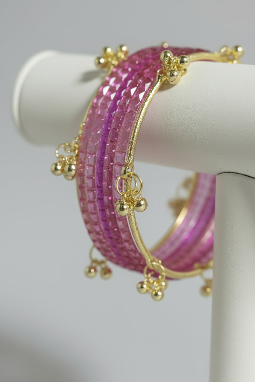 A set of pink beaded bangles with gold-toned metal borders and small dangling decorative charms, displayed on a wooden stick over black polka-dot wrapping paper.